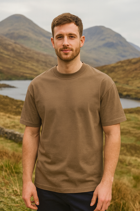 Man wearing a brown t-shirt standing in a scenic outdoor setting with mountains and a lake.